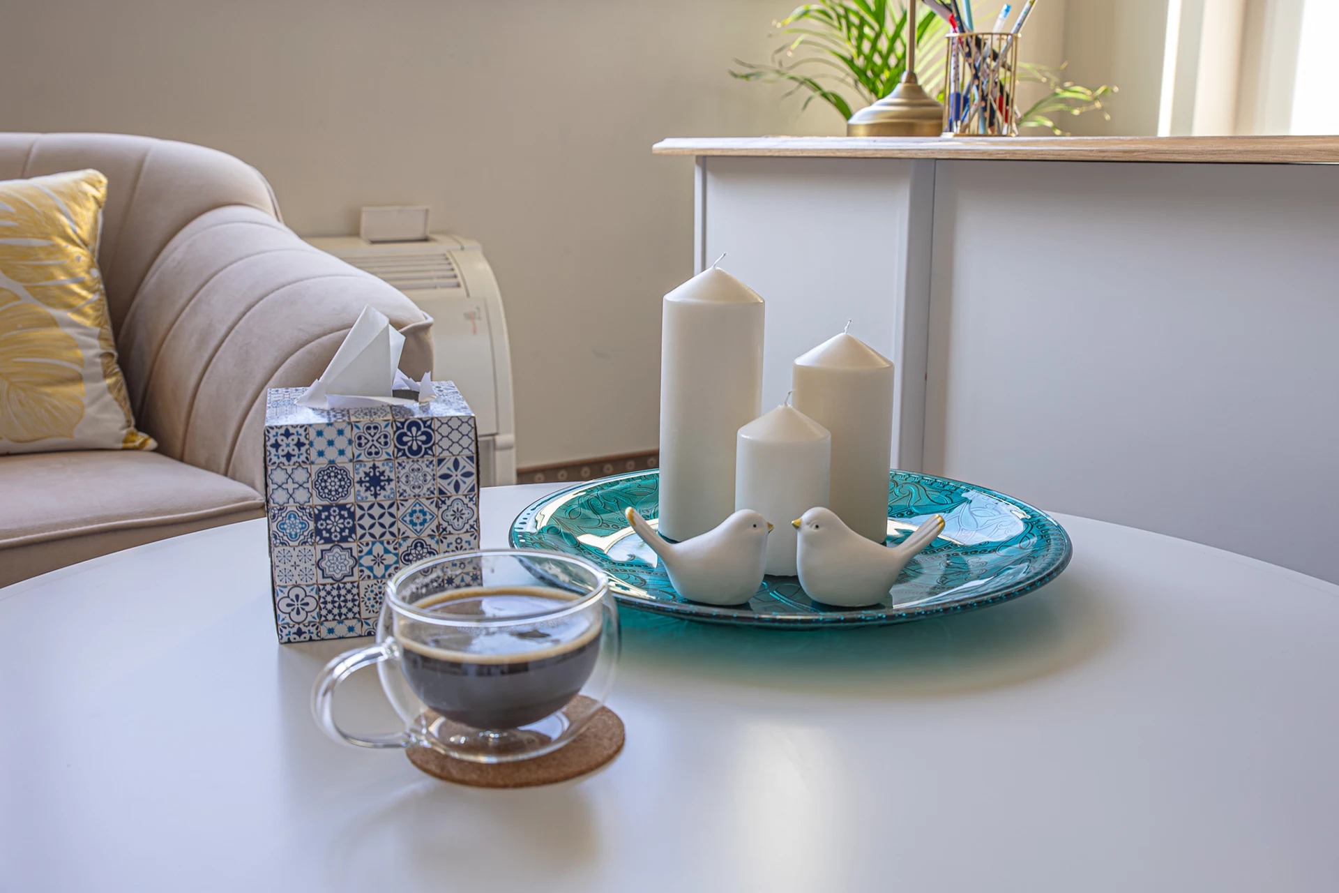 Jasne wnętrze z filiżanką kawy, pudełkiem chusteczek w niebieskie wzory, trzema białymi świecami i dwiema ceramicznymi figurkami ptaków na turkusowym talerzu.