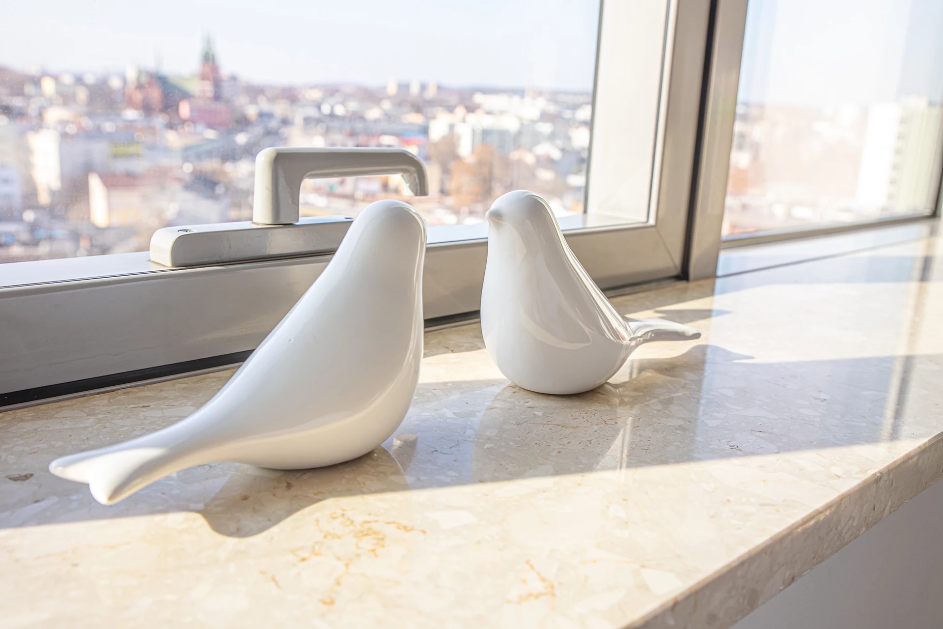 Dwie białe, ceramiczne figurki ptaków na marmurowym parapecie okiennym z widokiem na miasto i kościół w oddali, oświetlone jasnym światłem słonecznym.