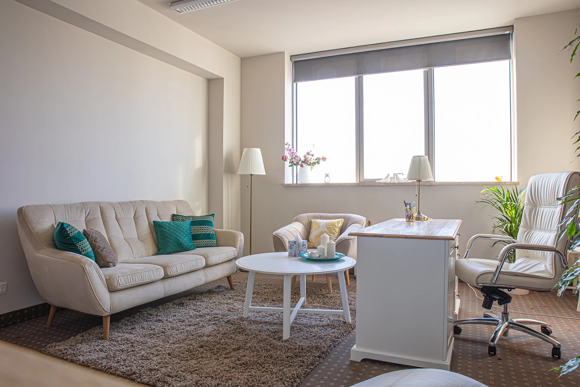 Jasny gabinet psychoterapeuty: beżowa sofa z turkusowymi poduszkami, biały stolik kawowy z dekoracjami, biurko z lampką, fotel biurowy, duże okno z roletą.