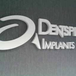 ADOS - Białe, trójwymiarowe litery 'DENTSPLY IMPLANTS' oraz stylizowane logo w kształcie spirali, zamontowane na ciemnoszarej, teksturowanej ścianie.