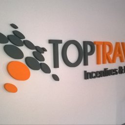 ADOS - Przestrzenny logotyp firmy TOP TRAVEL z hasłem Incentives & Holidays, wykonany z grubych liter i dynamicznych owalnych kształtów w szarości i pomarańczu, na białej ścianie.