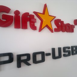 ADOS - Czerwone, trójwymiarowe litery 'GiftStar' z żółtą gwiazdą i czarne 'PRO-USB' zamontowane na białej ścianie, ujęcie z boku.