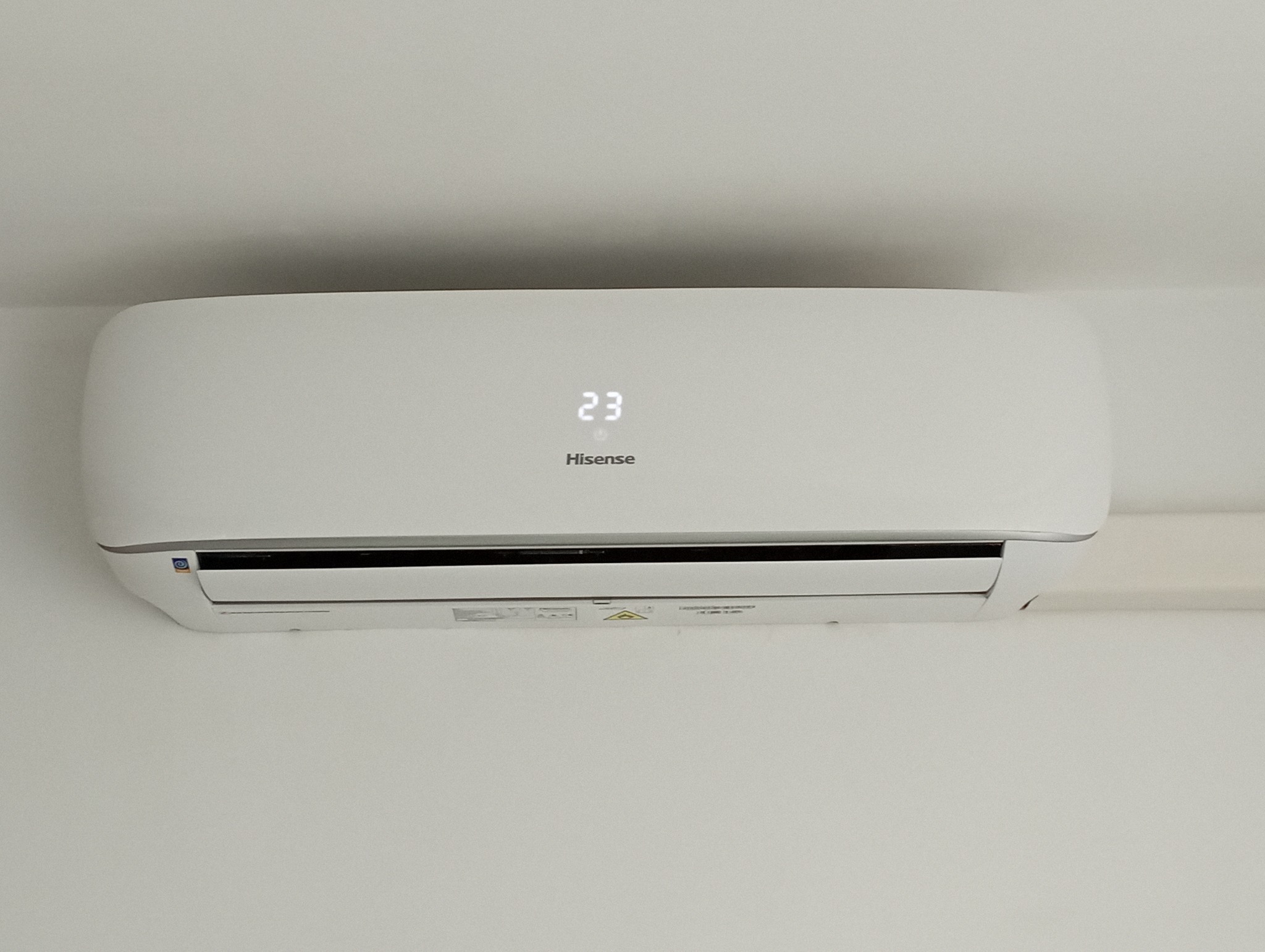 Biała jednostka klimatyzacji Hisense zamontowana na białej ścianie, wyświetlająca temperaturę 23 stopnie.