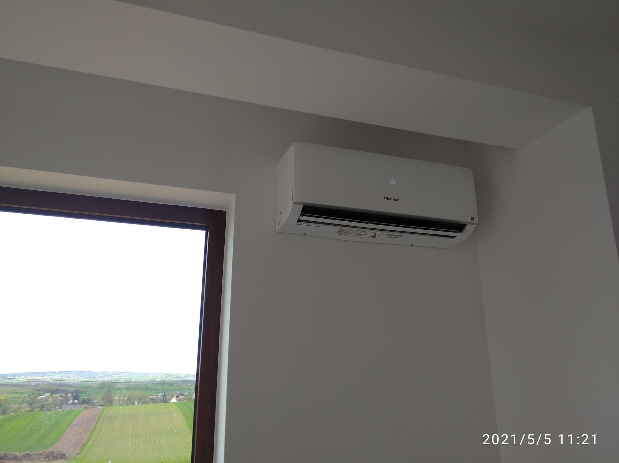 Biała jednostka wewnętrzna klimatyzatora Hisense zamontowana na ścianie w pomieszczeniu z widokiem na krajobraz przez okno.