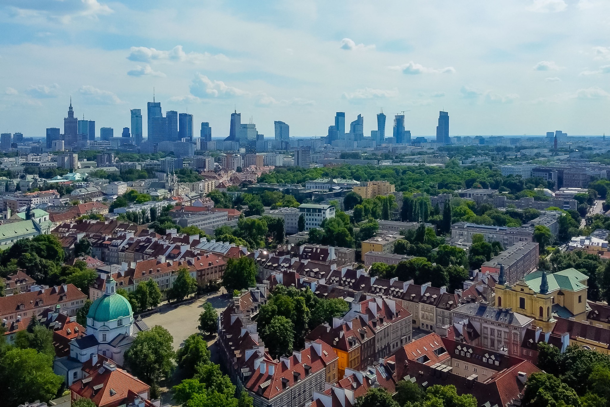 Panorama Warszawy z lotu ptaka, Stare Miasto z czerwonymi dachami i nowoczesne wieżowce w tle, błękitne niebo z chmurami, słoneczny dzień.