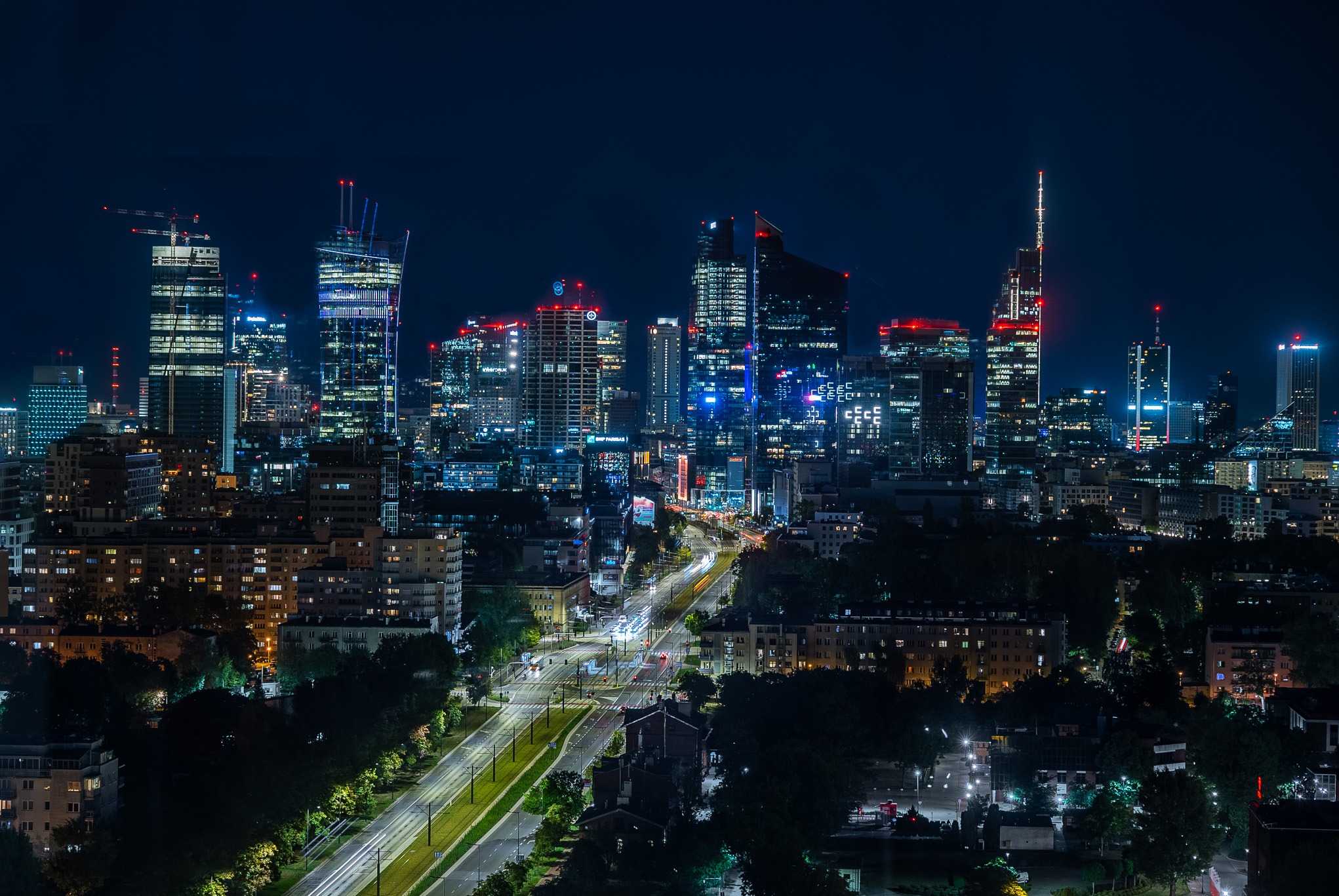Nocna panorama Warszawy z wieżowcami w neonowym blasku, widok z góry na oświetloną ulicę z rozmytymi światłami samochodów, zieleń drzew w dolnej części kadru.