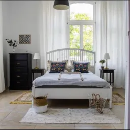 Jasna sypialnia z białym łóżkiem, wzorzystymi poduszkami i drewnianą tacą, widok przez otwarte drzwi z mosiężnymi klamkami, czarna komoda i lampki nocne, dywan z geometrycznym wzorem.