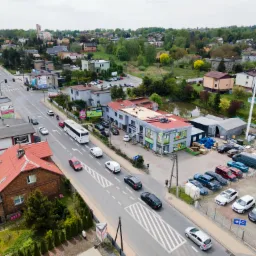 Widok z góry na budynek z panelami na dachu, duży parking i ulicę z przejeżdżającymi samochodami w tle zabudowa jednorodzinna i drzewa.