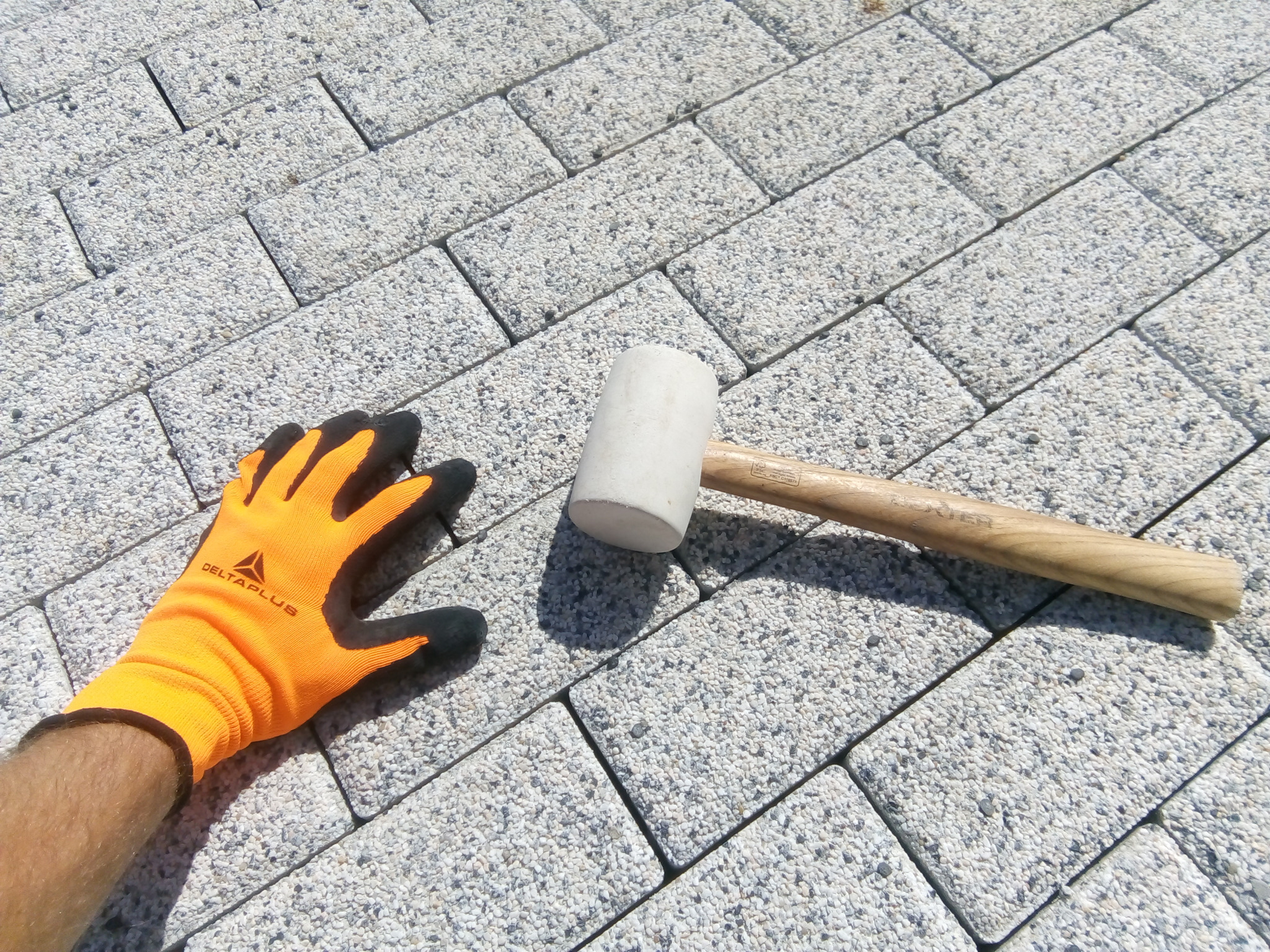 Świeżo ułożona, jasnoszara kostka brukowa z gumowym młotkiem i rękawicą roboczą Delta Plus, widok z góry.