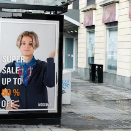Plakat reklamowy w wiacie przystankowej z młodym mężczyzną i napisem 'SUPER SALE UP TO 50% OFF' na tle ulicy z budynkami i koszami na śmieci.