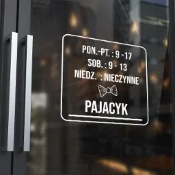 Naklejka z godzinami otwarcia i logo 'Pajacyk' na szklanych drzwiach wejściowych do lokalu.