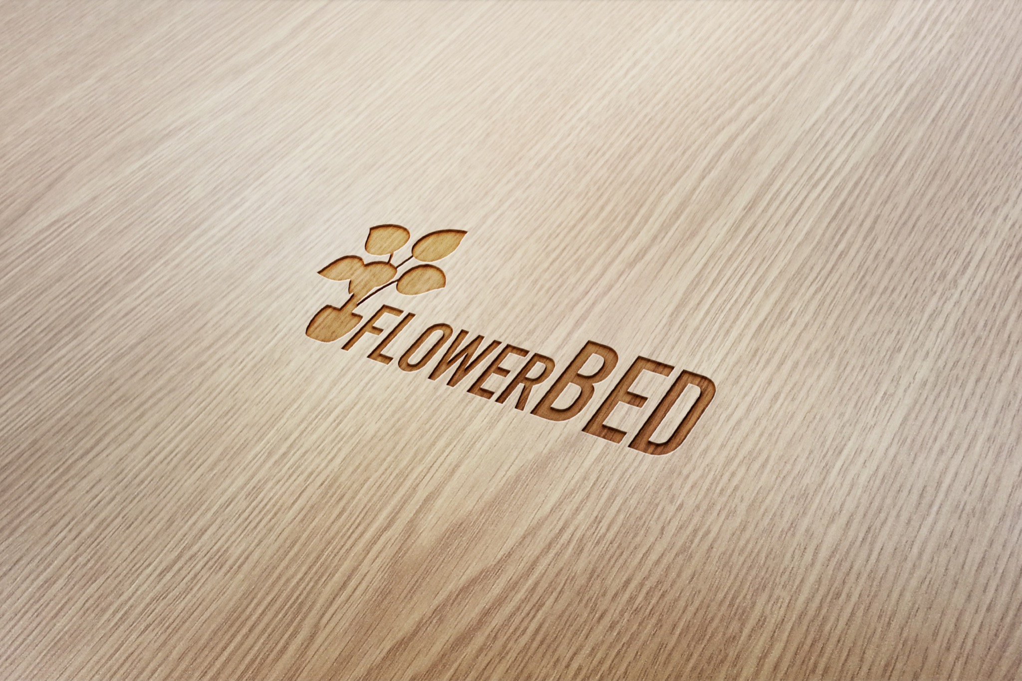 Logo 'FLOWERBED' z motywem roślinnym, wygrawerowane na jasnym, drewnianym tle z widoczną strukturą drewna, ustawione pod kątem.