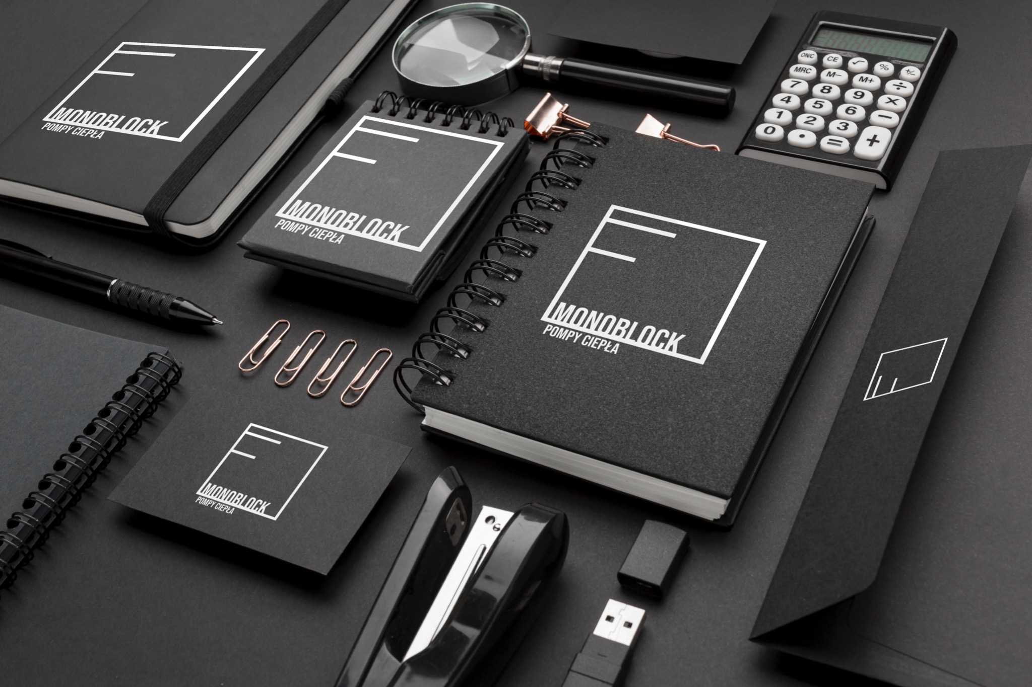 Zestaw materiałów biurowych w czerni: notatniki, wizytówka, długopis, spinacze, zszywacz, kalkulator i lupa, z logo firmy 'Monoblock Pompy Ciepła' na każdym elemencie, ułożone na czarnym tle.