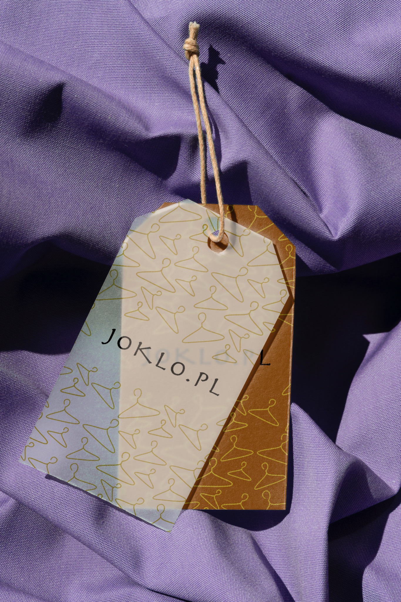 Metka odzieżowa Joklo.pl na fioletowym tle materiału koszulowego, z motywem wieszaków i sznurkowym zawiesiem.
