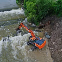 Pomarańczowa koparka gąsienicowa w korycie rzeki, prace ziemne przy brzegu wzmocnionym palisadą.