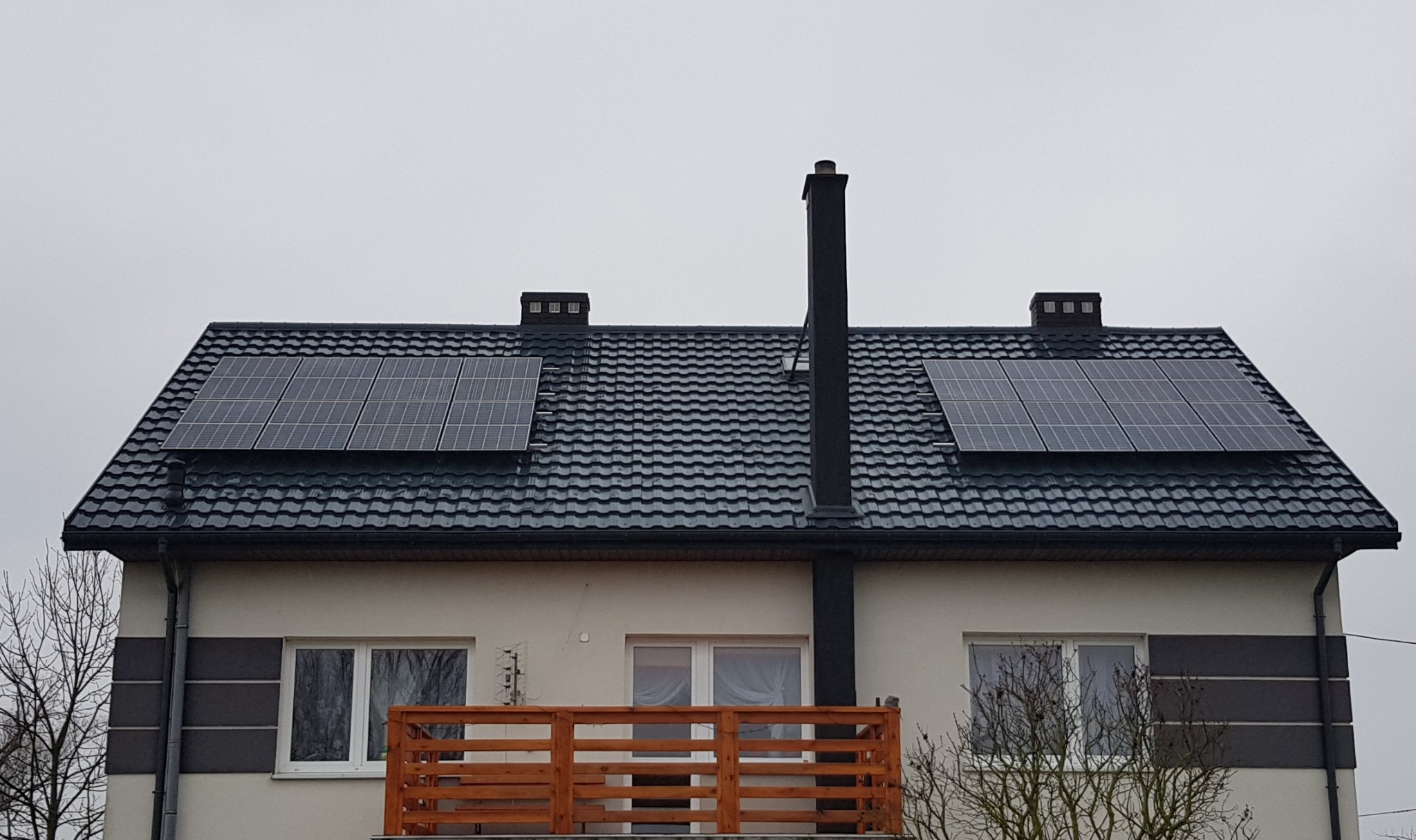 Dwa panele słoneczne zamontowane na dachu dwupiętrowego domu z ciemną dachówką, widoczne kominy i balkon z drewnianą balustradą, pochmurne niebo w tle.