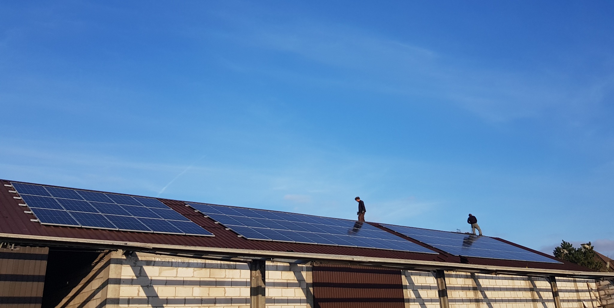 Dwaj instalatorzy na dachu budynku gospodarczego montują panele słoneczne; widoczny jasny mur z pustaków i błękitne niebo.