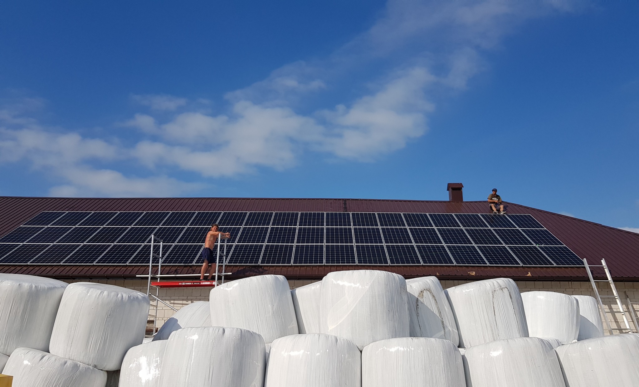 Montaż paneli słonecznych na dachu budynku gospodarczego; widoczni dwaj mężczyźni na dachu, jeden na rusztowaniu, drugi siedzący na kalenicy; w tle stogi białych bel siana.