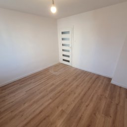 Jan Pawlicki - Pusty pokój po remoncie: jasne ściany, panele podłogowe w odcieniach brązu, białe drzwi z przeszkleniami i wisząca żarówka bez klosza.