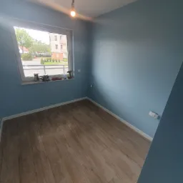 Pomalowane na niebiesko wnętrze pokoju z drewnopodobną podłogą, z oknem na którym stoją puszki z farbą i wiertarka, oraz wisząca żarówka na kablu.