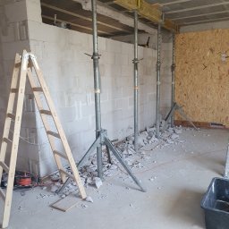 Jan Pawlicki - Wnętrze w trakcie prac murarskich: ściana z betonu komórkowego z otworem wzmocniona stemplami budowlanymi, drewniana drabina oparta o ścianę, gruz na podłodze.