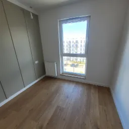 Pusty pokój z panelami podłogowymi w odcieniach brązu, grzejnikiem pod oknem z widokiem na parking i budynek mieszkalny, oraz oliwkową ścianą z dekoracyjnymi panelami i gniazdkiem elektrycznym.