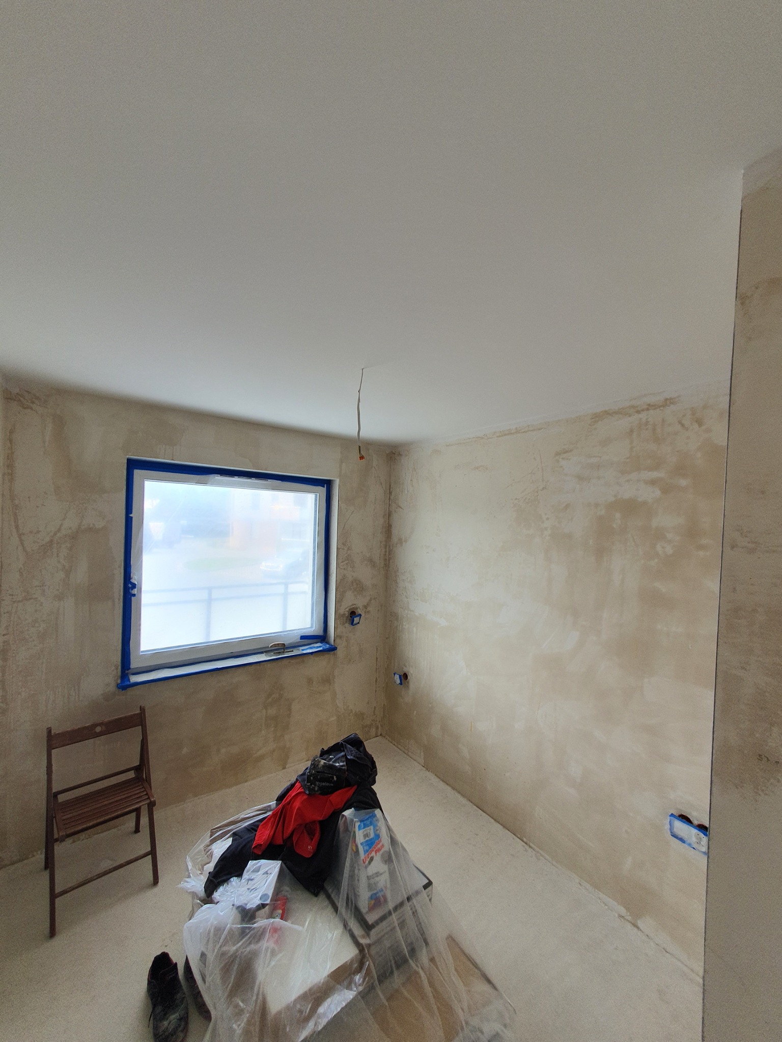 Pokój w trakcie szpachlowania ścian, okno zabezpieczone taśmą malarską, widoczne narzędzia, ubrania robocze i krzesło.