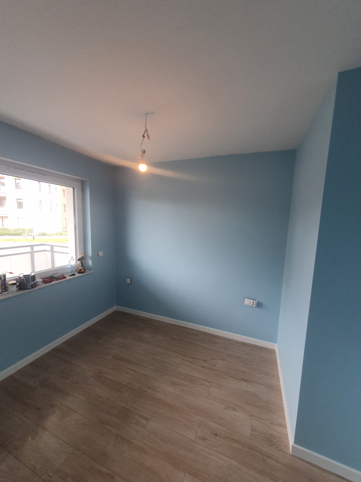 Pomalowane na błękitno wnętrze pokoju z drewnianą podłogą i oknem, na parapecie narzędzia i puszki farby, surowa żarówka zwisa z sufitu.