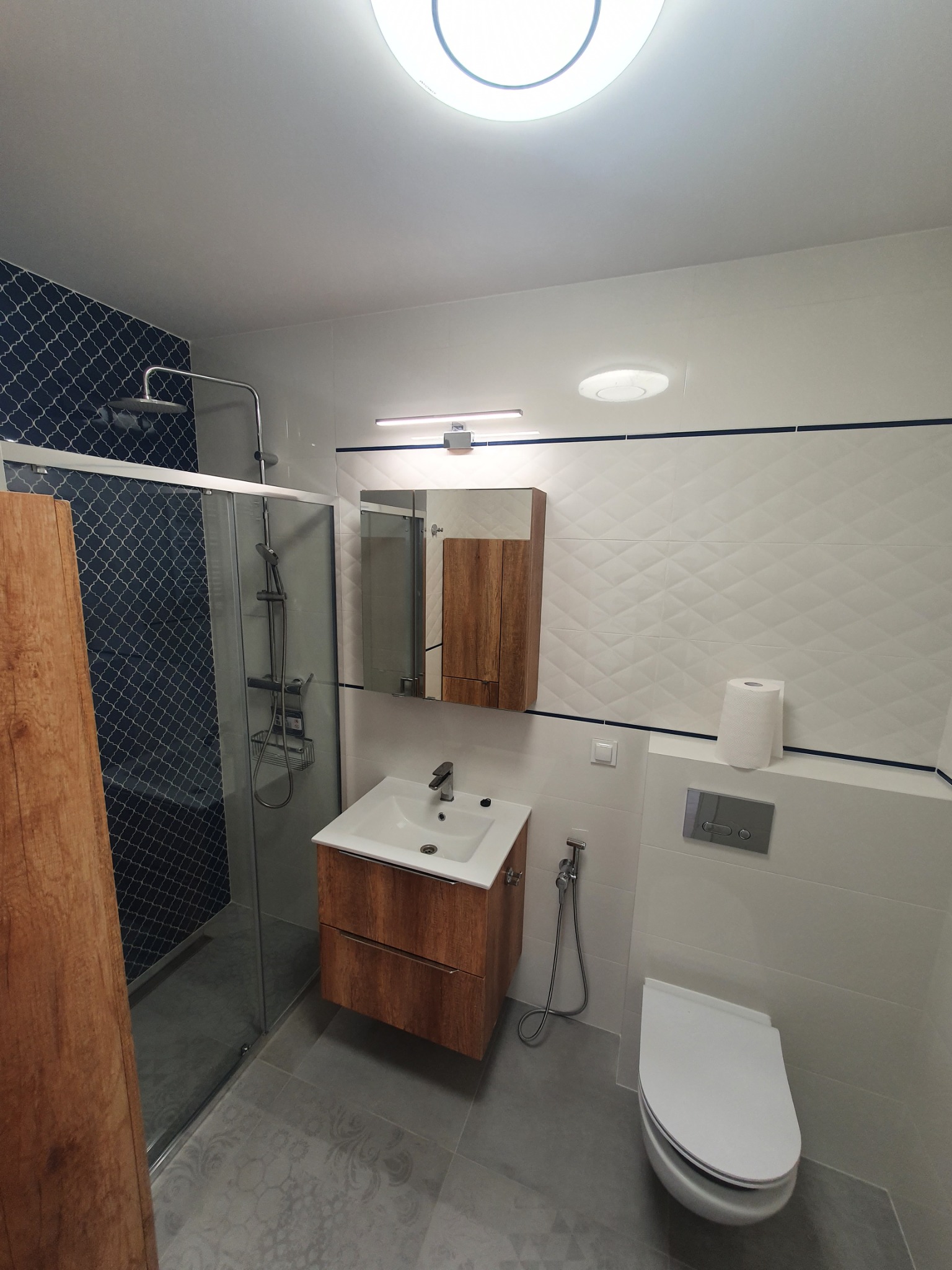 Nowoczesna łazienka z kabiną prysznicową z deszczownicą, umywalką z szafką z drewnopodobnym fornirem, toaletą podwieszaną i białymi płytkami ze wzorem rombów.