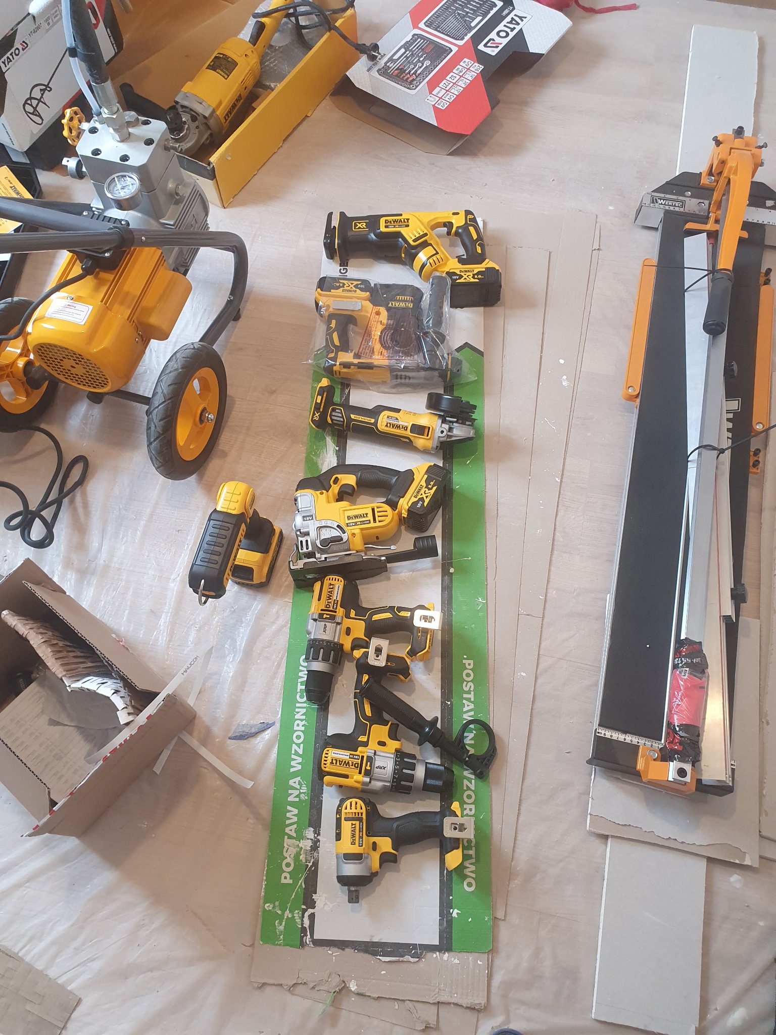 Zestaw narzędzi DeWalt, w tym przecinarka do płytek, szlifierka kątowa, wiertarki i inne, ułożonych na podłodze z kartonami i folią ochronną, sugerujących przygotowanie do prac wykończeniowych.