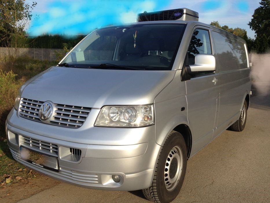 Srebrny Volkswagen Transporter z agregatem chłodniczym Carrier na dachu, zaparkowany na asfalcie, widok z przodu na tle zieleni.