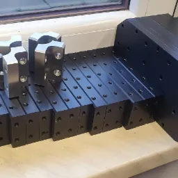 Czarne, precyzyjnie obrobione elementy z otworami, ułożone rzędami na jasnym blacie, z trzema srebrnymi detalami o skomplikowanym kształcie na wierzchu, widoczne ślady obróbki.