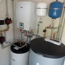 Instalacja grzewcza z białym kotłem gazowym, dwoma zbiornikami (białym i niebieskim) oraz dwoma zasobnikami ciepłej wody (białym i szarym) marki Buderus i Kospel, widoczne rury miedziane i zawory...