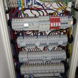 Wewnętrzny widok rozdzielnicy elektrycznej z licznymi przewodami, bezpiecznikami i zabezpieczeniami przepięciowymi, uporządkowanymi na szynach montażowych w białej obudowie.