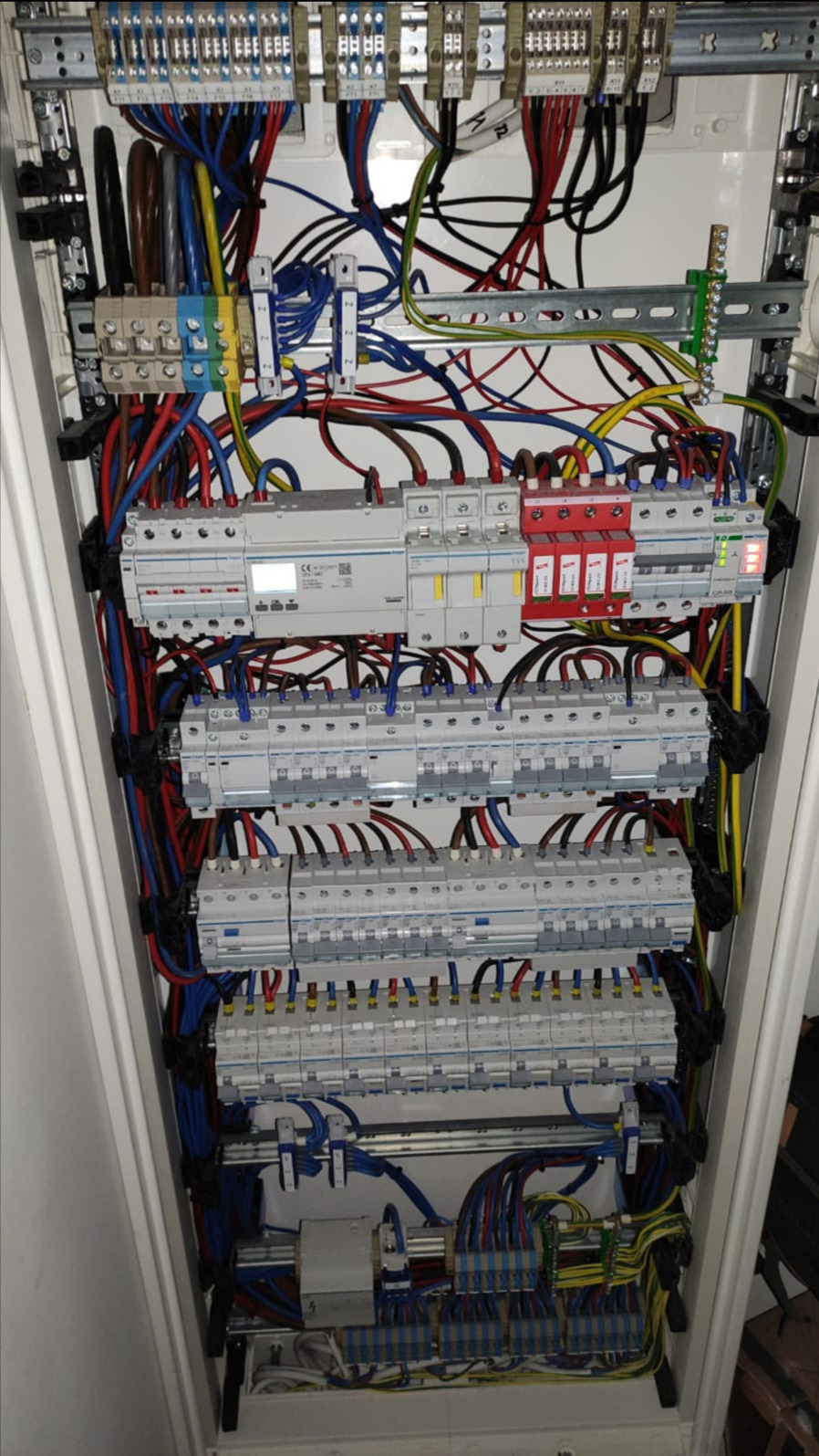 Wewnętrzny widok rozdzielnicy elektrycznej z licznymi przewodami, bezpiecznikami i zabezpieczeniami przepięciowymi, uporządkowanymi na szynach montażowych w białej obudowie.