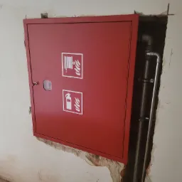 Czerwona szafka hydrantowa z piktogramami węża strażackiego i gaśnicy, zamontowana w ścianie z widocznymi instalacjami hydraulicznymi w nieotynkowanym otworze.