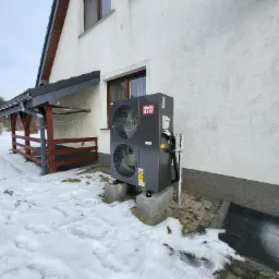 Szara pompa ciepła z dwoma wentylatorami, zamontowana na betonowych podstawach przy białym budynku w zimowej scenerii, widoczne rury i logo producenta.