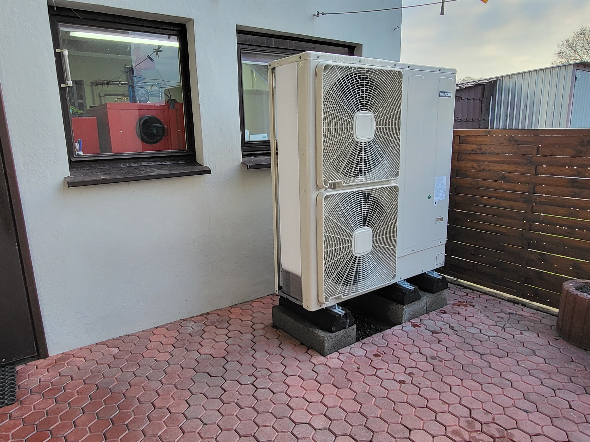 Zewnętrzna jednostka pompy ciepła Hitachi, zamontowana na betonowych blokach na czerwonej kostce brukowej, widok z boku budynku z oknem, za nią drewniany płot i blaszany garaż.
