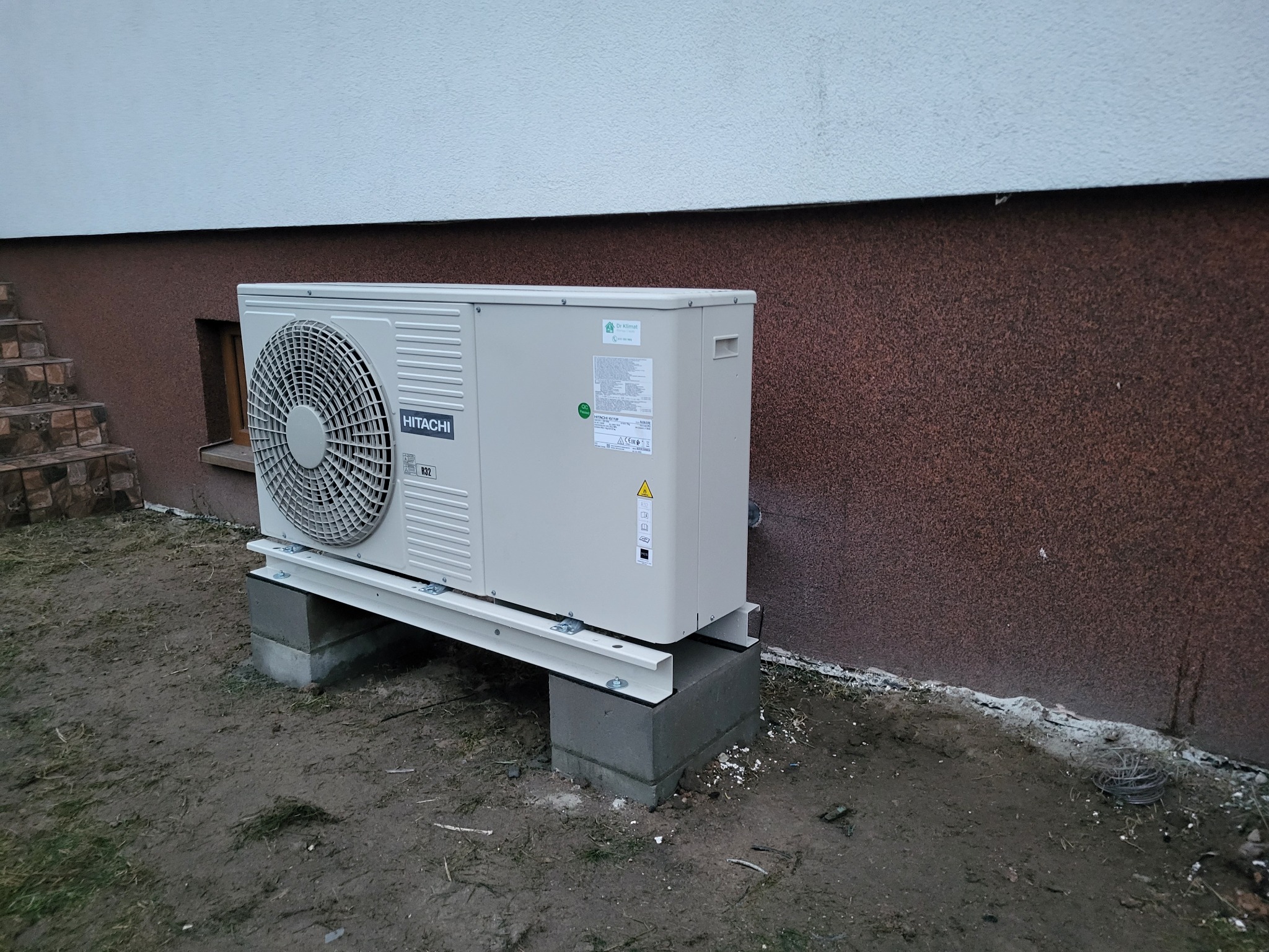 Zewnętrzna jednostka klimatyzacji Hitachi zamontowana na betonowych bloczkach i metalowej konstrukcji, przy ścianie budynku z widocznymi schodami i ziemistym podłożem.