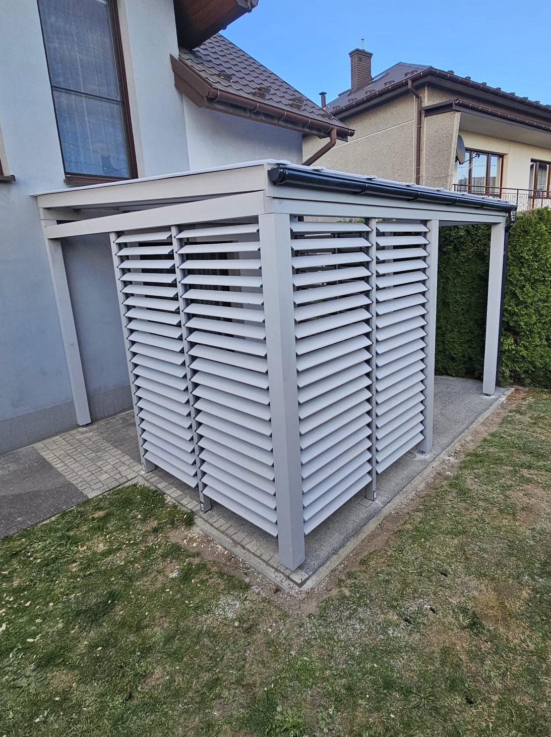 Nowoczesna, szara pergola lamelowa przy budynku z widocznym systemem odprowadzania wody. Elementy betonowe wokół podstawy, trawnik w tle.