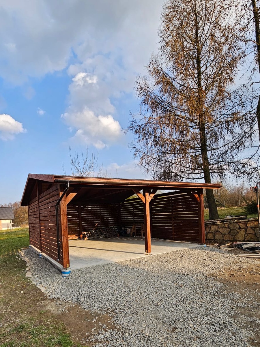 Drewniana wiata garażowa z ażurowymi ścianami bocznymi, umiejscowiona na żwirowym podłożu. Widoczne elementy konstrukcyjne i brązowe wykończenie drewna.