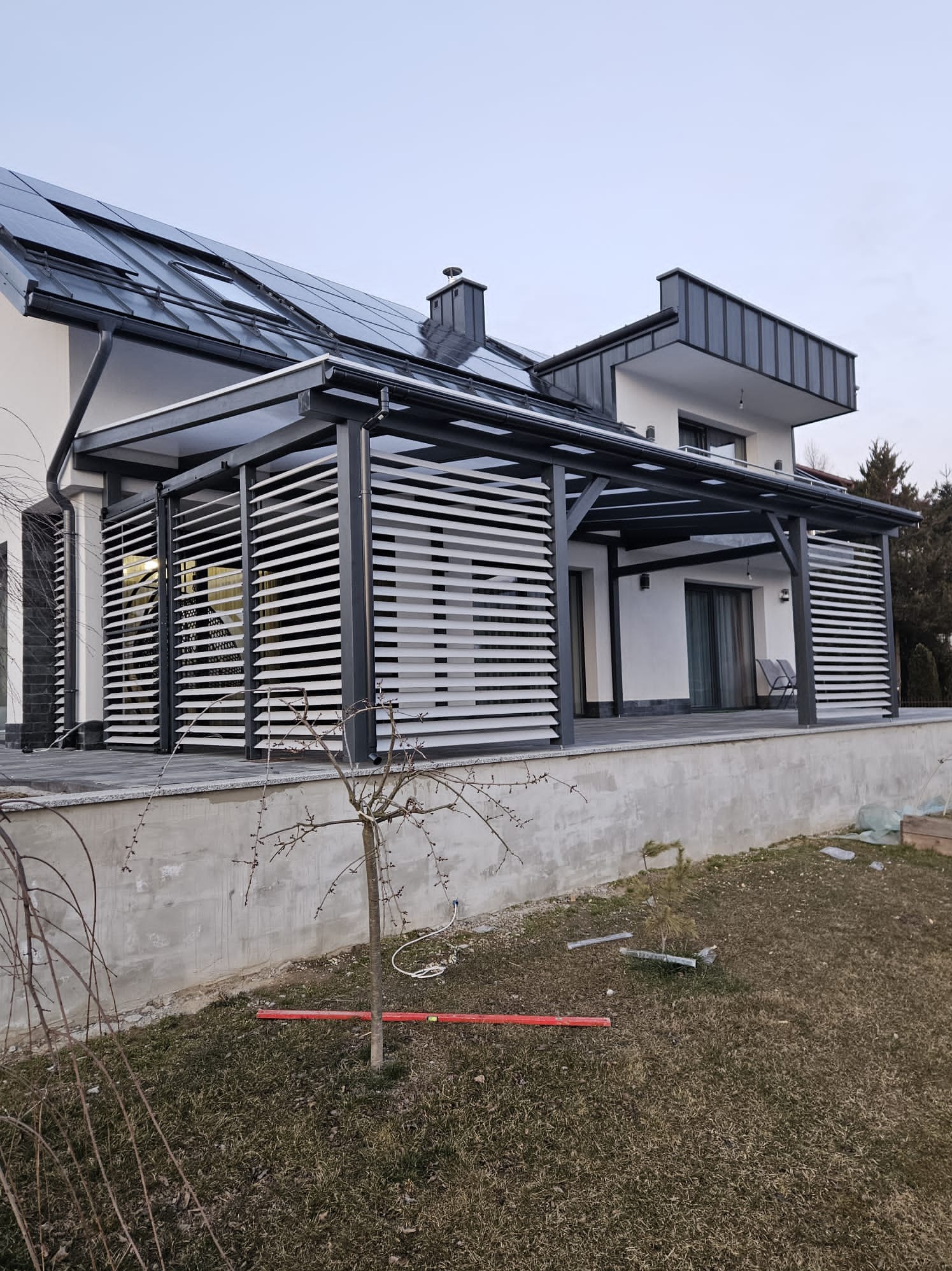Nowoczesna, szara pergola z lamelami przy domu z panelami słonecznymi. Widoczny taras i elementy ogrodu. Minimalistyczny design.