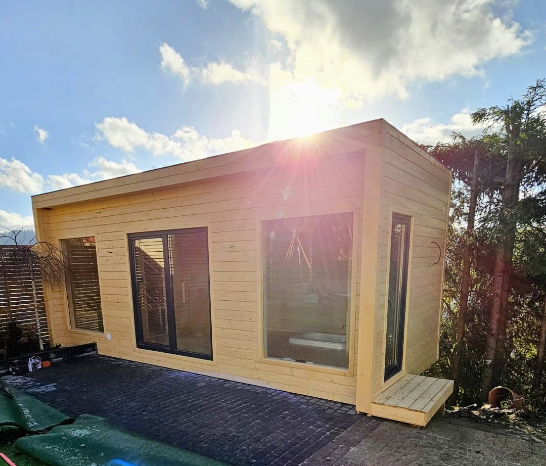 Nowoczesna, drewniana altana z dużymi oknami, oświetlona promieniami słońca. Ułożona kostka brukowa i fragment trawy w rolkach. Widoczne drzewa w tle.