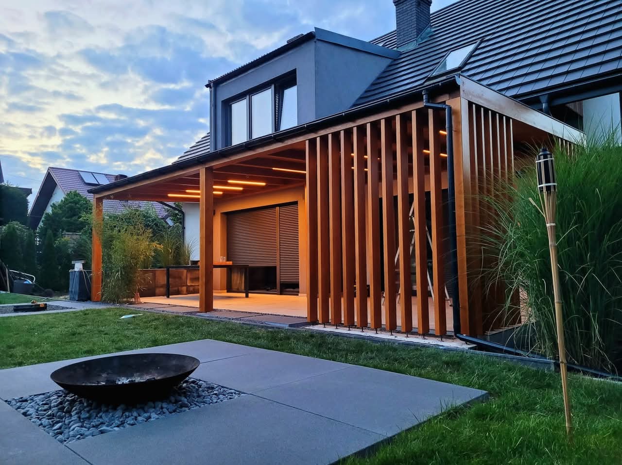 Nowoczesna pergola z drewnianymi lamelami i oświetleniem LED, przylegająca do domu z ciemną dachówką. W tle trawnik i palenisko.