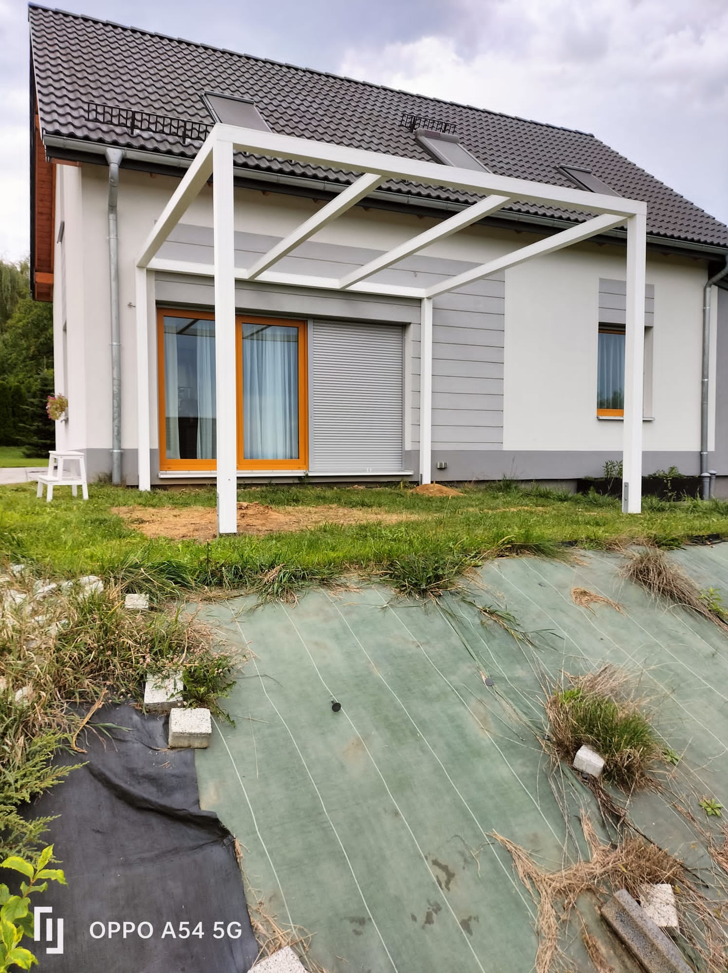 Biała, nowoczesna pergola z płaskim dachem, przylegająca do jasnego domu z brązowymi ramami okiennymi. Widok na ogród z zieloną tkaniną i kamieniami.