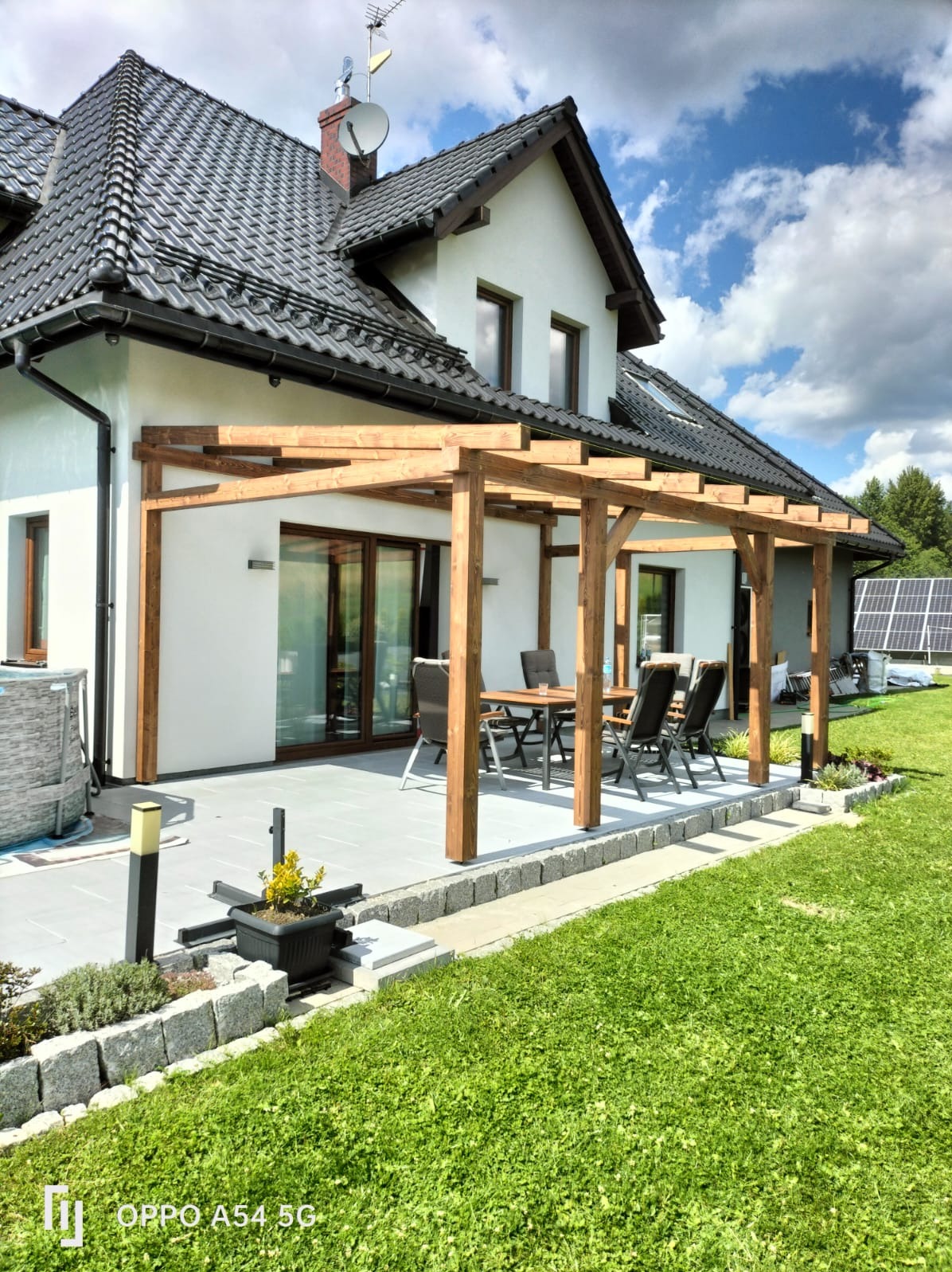 Drewniana pergola przy domu z szarym tarasem i zestawem mebli ogrodowych. Widoczne panele słoneczne w tle, zieleń trawnika i błękitne niebo z chmurami.