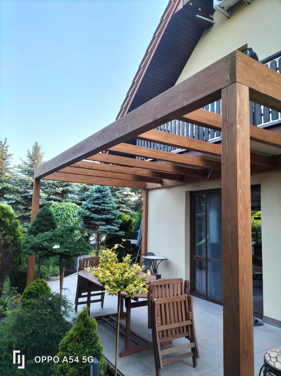 Brązowa pergola z drewna na tarasie przy domu, otoczona zielenią. Widoczne meble ogrodowe i rośliny ozdobne, tworzące przytulną przestrzeń.