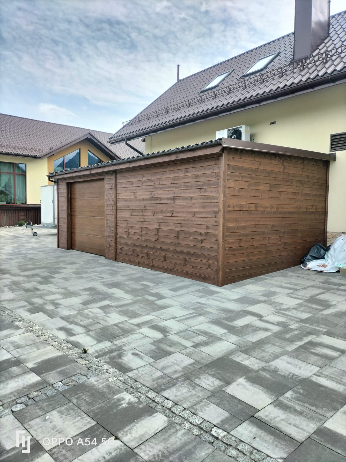 Drewniana zabudowa z przesuwnymi drzwiami, przylegająca do budynku z brązowym dachem. Wykonana z desek w ciepłym odcieniu brązu, na szarej kostce brukowej.