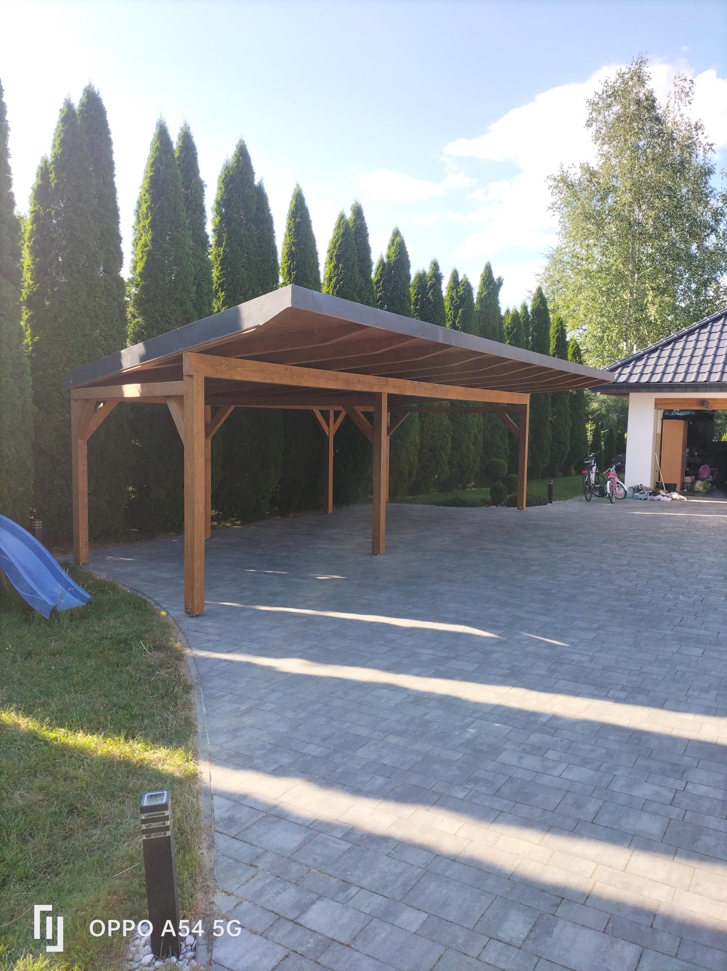 Drewniana pergola z płaskim dachem na posesji. W tle wysokie tuje i budynek garażowy. Na podjeździe rowery i zabawki.