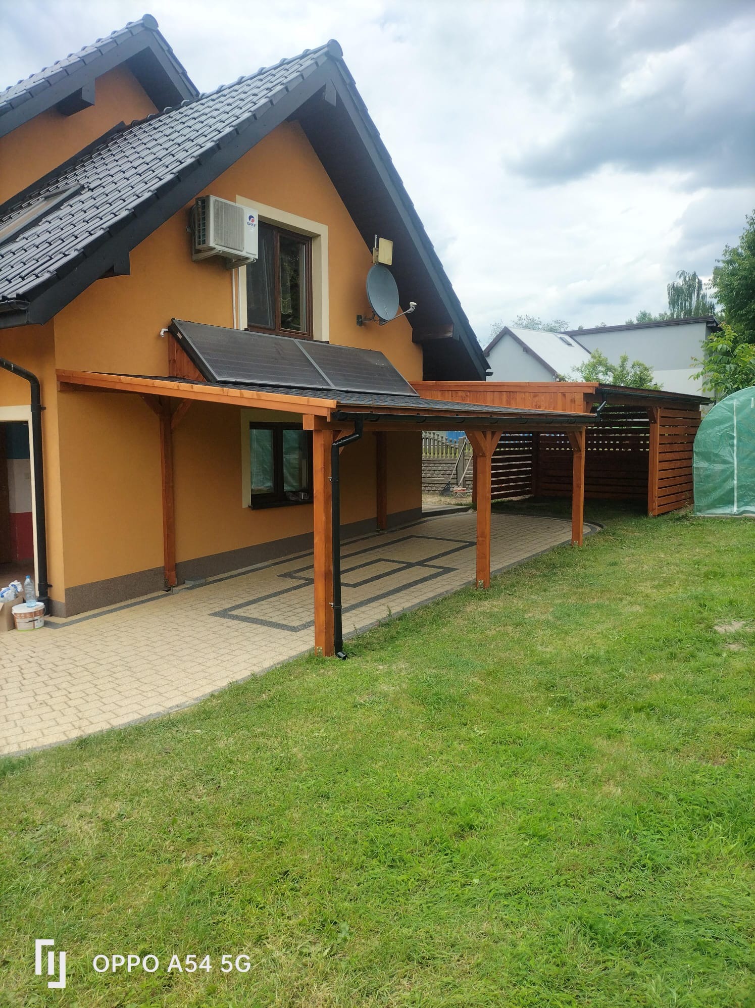 Zadaszenie tarasu z drewna przy domu z panelami słonecznymi na dachu. Widoczny fragment ogrodu i ażurowa konstrukcja w tle.